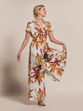 Beige solid-color jumpsuit with floral tan colors, shown on model standing on one leg, side view, looking sideways.