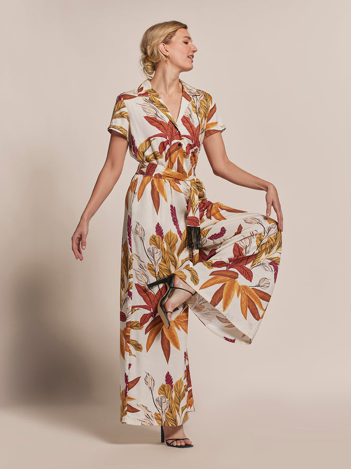 Beige solid-color jumpsuit with floral tan colors, shown on model standing on one leg, side view, looking sideways.