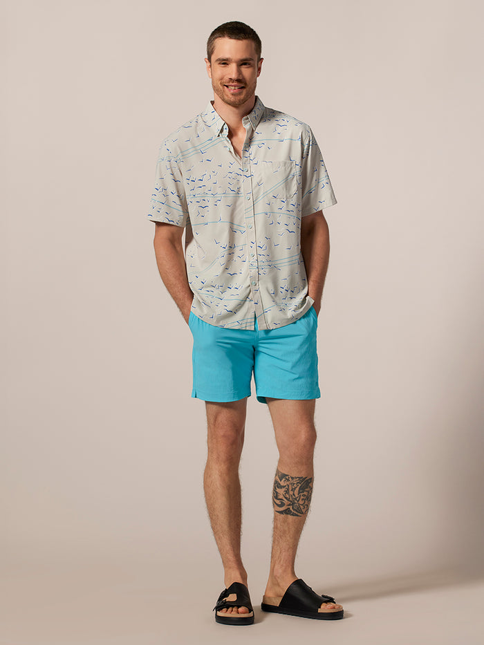 Light gray short-sleeve button-up shirt with blue bird print solid short-sleeve Tradewind Technology® shirt and blue shorts, front view.
