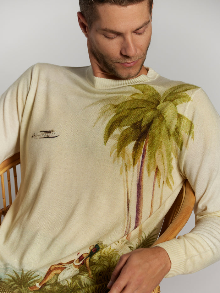 Cream and tan tropical scene-print long-sleeve sweater, seated front view on wooden chair.