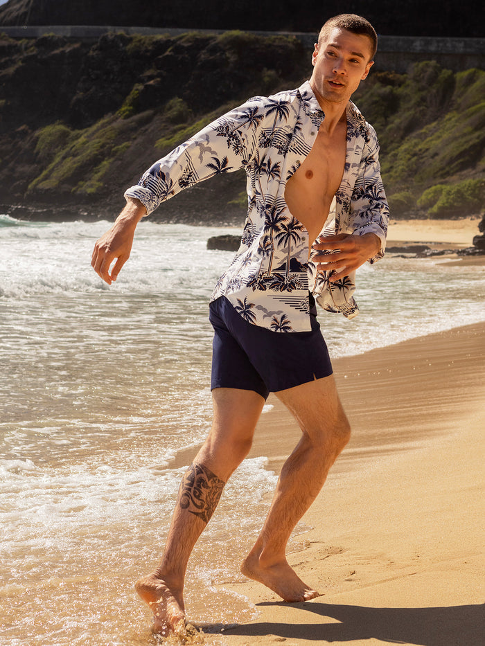 White with blue and brown palm print, long sleeves long-sleeve performance shirt and navy shorts, model walking on beach, side view.