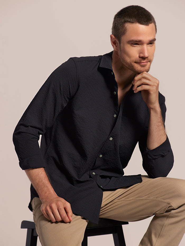 Black solid long-sleeve shirt with chest pocket and tan pants, seated side view.