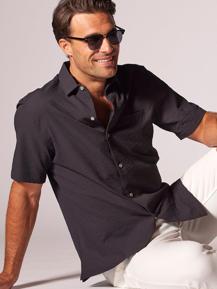 Black tropical-print short-sleeve shirt worn by man lounging against light background, front view.