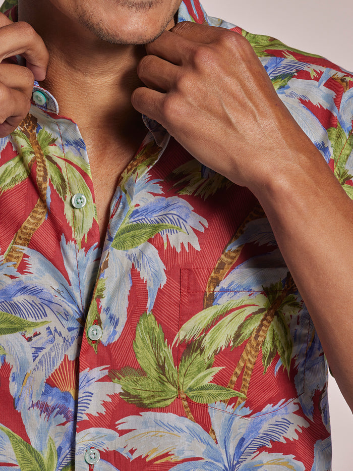 Red short-sleeve shirt with blue, green, and white palm leaf print, torso and hands adjusting shirt front view.