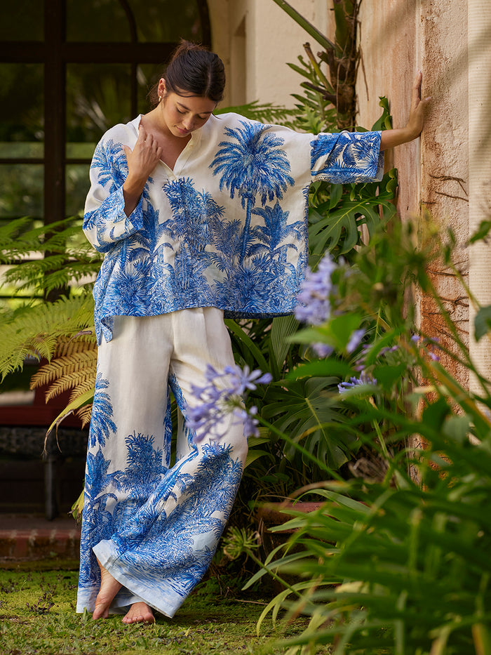 Barefoot woman in blue floral-print top stands among green plants and flowers, front view.