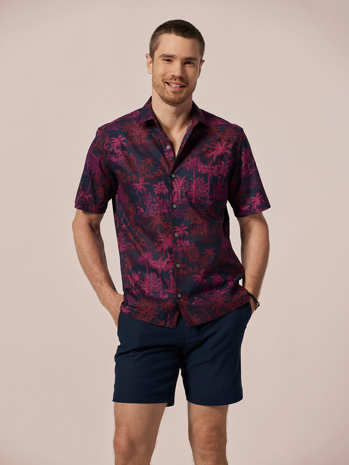 Red palm tree print short-sleeve shirt and navy shorts; man smiling, full front view.