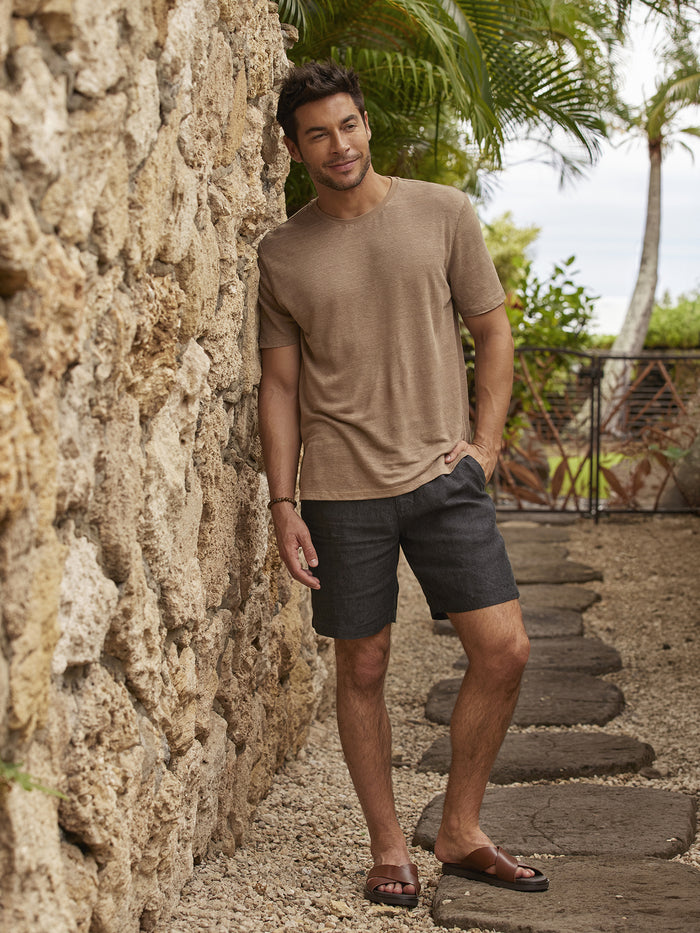Tan solid linen short-sleeve T-shirt, side view, man leaning on stone wall outdoors (images 1 & 2).
