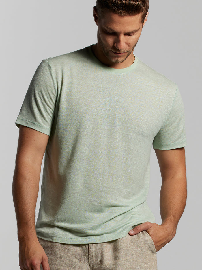 Light green graphic-print Vacay T-shirt, beige pants, man looking down with hand in pocket, front view.