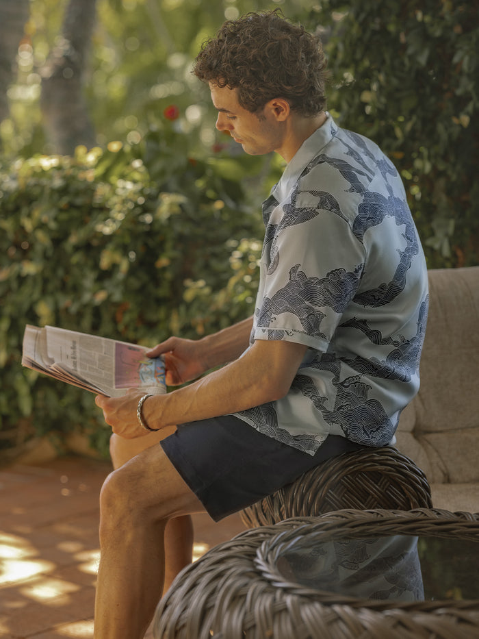 Light blue Water Wheel print silk shirt and shorts, worn by a seated model in a front view on a wicker bench.