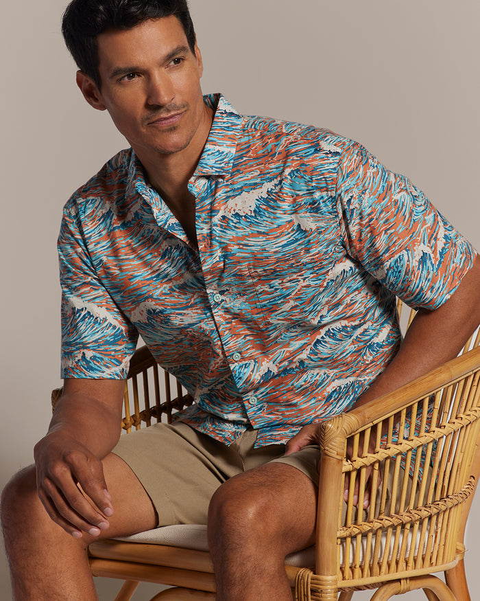 White/blue and orange wave abstract-print cotton short-sleeve shirt with khaki shorts, seated side view.